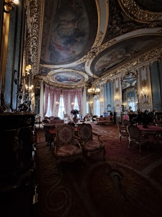 A lavish and opulent room with ornate ceiling paintings and intricate gold detailing throughout. The room features luxurious red and gold upholstered chairs and sofas, with elegant chandeliers hanging from the ceiling. Large draped windows allow natural light to illuminate the rich interior, creating a warm and sophisticated atmosphere.