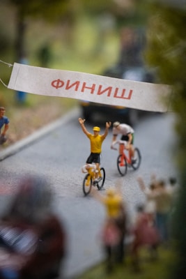 Toy cyclists ride towards a finish line marked by a banner with text written in Cyrillic. The scene appears to be a miniature model, featuring two cyclists with one raising their arms in victory. The background includes blurred spectators and trees, emphasizing a sense of motion and scale.