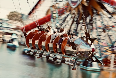 A group of people are riding a vintage-style swing ride shaped like a rocket ship, with a ferris wheel visible in the background. The ride is in motion over a body of water, adding a sense of excitement and thrill to the scene.