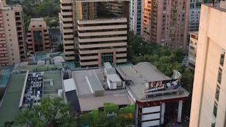 Aerial view of an urban area featuring multiple high-rise buildings with a mix of modern architectural styles. The rooftops display various mechanical installations, and trees can be seen lining the streets below. A large billboard on a building advertises a local business or product.