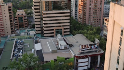 Aerial view of an urban area featuring multiple high-rise buildings with a mix of modern architectural styles. The rooftops display various mechanical installations, and trees can be seen lining the streets below. A large billboard on a building advertises a local business or product.