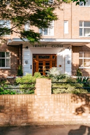 A brick building entrance with the sign 'Trinity Close' above the door. The facade features large windows and a white-framed doorway with a wooden door. In front of the building, there is a well-maintained garden with various green plants and a low brick wall bordering the sidewalk. Sunlight casts shadows of leaves across the wall and pathway.