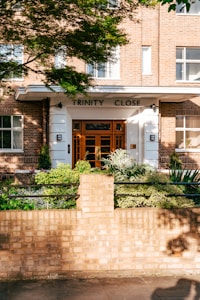 A brick building entrance with the sign 'Trinity Close' above the door. The facade features large windows and a white-framed doorway with a wooden door. In front of the building, there is a well-maintained garden with various green plants and a low brick wall bordering the sidewalk. Sunlight casts shadows of leaves across the wall and pathway.