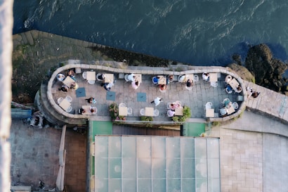 Aerial view of an outdoor seating area located beside a body of water. The space is occupied by several small tables with people sitting on white chairs. The seating area is enclosed by a stone wall and features potted plants. The water body is on the right, while the stone pavement and a glass-roofed structure are visible on the left.