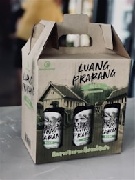 A cardboard carrier box contains several cans of beer labeled 'Luang Prabang.' The packaging is adorned with illustrations of traditional buildings, likely representing the heritage of Luang Prabang, a city in Laos. The carrier has a handle integrated into its design for easy transport.