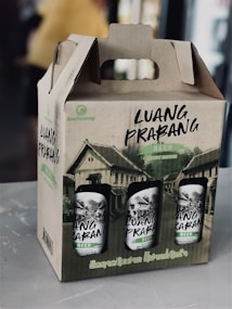 A cardboard carrier box contains several cans of beer labeled 'Luang Prabang.' The packaging is adorned with illustrations of traditional buildings, likely representing the heritage of Luang Prabang, a city in Laos. The carrier has a handle integrated into its design for easy transport.