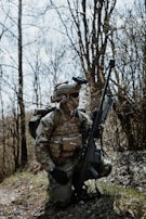 A person wearing camouflage military gear, including a helmet and tactical vest, kneels on a grassy area amidst bare trees. The individual holds a large rifle and carries a backpack, appearing to be in a forest environment.