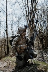 A person wearing camouflage military gear, including a helmet and tactical vest, kneels on a grassy area amidst bare trees. The individual holds a large rifle and carries a backpack, appearing to be in a forest environment.
