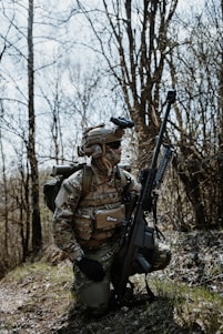 A person wearing camouflage military gear, including a helmet and tactical vest, kneels on a grassy area amidst bare trees. The individual holds a large rifle and carries a backpack, appearing to be in a forest environment.