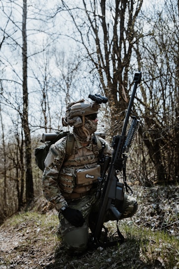 A person wearing camouflage military gear, including a helmet and tactical vest, kneels on a grassy area amidst bare trees. The individual holds a large rifle and carries a backpack, appearing to be in a forest environment.