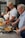 Three people are cooking together in a kitchen. An elderly man is stirring vegetables in a skillet on the stove, while a man and woman are smiling and preparing more vegetables on a cutting board.