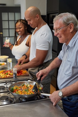 Three people are cooking together in a kitchen. An elderly man is stirring vegetables in a skillet on the stove, while a man and woman are smiling and preparing more vegetables on a cutting board.