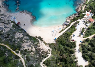An aerial view of a serene beach featuring clear turquoise waters and a sandy shoreline. There are several rows of sunbeds with umbrellas arranged neatly on the sand. The surrounding area includes rocky outcrops and lush green vegetation. A few buildings and a winding road with parked cars are visible on the right.