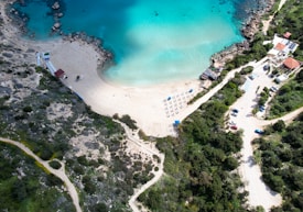An aerial view of a serene beach featuring clear turquoise waters and a sandy shoreline. There are several rows of sunbeds with umbrellas arranged neatly on the sand. The surrounding area includes rocky outcrops and lush green vegetation. A few buildings and a winding road with parked cars are visible on the right.