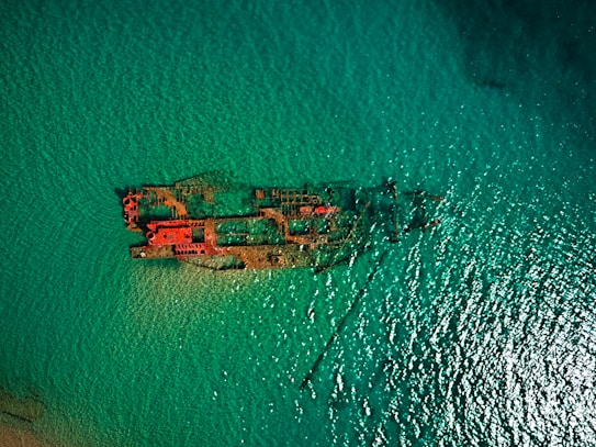 An aerial view of a partially submerged shipwreck in clear turquoise waters. The rusted metal structure of the ship contrasts with the vibrant color of the ocean, creating an intriguing and mysterious atmosphere.