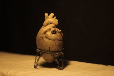 A sculpture resembling a human heart is placed on a metal stand made of twisted wires. The lighting casts a warm, soft glow on the sculpture, set against a dark background, enhancing its textures and details.