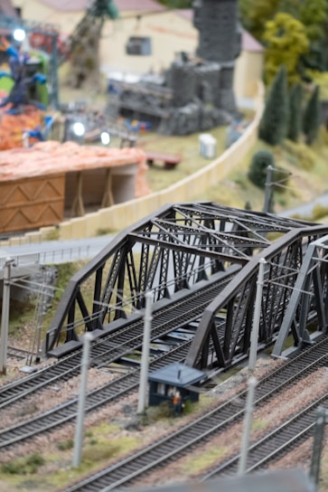 A detailed model train set scene featuring a steel bridge over multiple train tracks. In the background are miniature houses, trees, and structures, suggesting a small town or village setting. The scene includes fine details such as poles, tracks, and a small human figure.