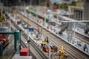 A detailed miniature railway model depicting multiple train tracks, surrounded by tiny figures and constructed structures. There are model trains on the tracks, with various small people figures engaged in different activities around the scene. The intricate setup includes overhead cables, model buildings, and construction equipment.
