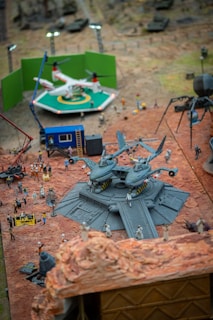 A detailed miniature scene features a set of aircraft models on a raised platform surrounded by small figurines of people. The setting includes a green screen backdrop behind a helicopter model on a landing pad. Lights and other set equipment, such as cranes and trailers, are present, creating the appearance of a film set.
