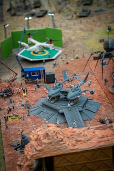 A detailed miniature scene features a set of aircraft models on a raised platform surrounded by small figurines of people. The setting includes a green screen backdrop behind a helicopter model on a landing pad. Lights and other set equipment, such as cranes and trailers, are present, creating the appearance of a film set.
