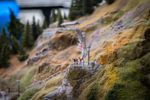 A miniature scene depicts a rocky landscape with small human figures walking along a path. The path is bordered by railings and dotted with lampposts. In the background, coniferous trees are visible, adding depth to the scene.