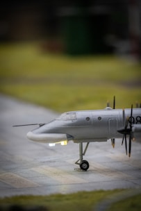 A model airplane sits on a flat surface with its landing gear down. The aircraft is painted in a metallic gray color, and several propellers are visible along its side. Small, glowing lights are present near the nose and underbelly, giving the model a realistic appearance. The background is softly blurred, focusing attention on the plane.