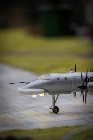 A model airplane sits on a flat surface with its landing gear down. The aircraft is painted in a metallic gray color, and several propellers are visible along its side. Small, glowing lights are present near the nose and underbelly, giving the model a realistic appearance. The background is softly blurred, focusing attention on the plane.