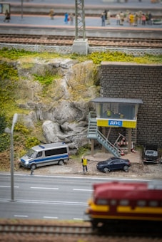 A detailed miniature scene depicting a roadside scenario with a model police station or checkpoint, including a sign with Cyrillic text. Two vehicles, a van and a car, are parked nearby, with one figure dressed in a yellow vest standing beside them. A red train is slightly blurred in the foreground, indicating proximity to a railway line. The setting includes rocky terrain with greenery and railway tracks in the background, with miniature figures representing people at a train station.