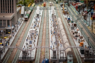 A detailed model of a bustling train station with multiple platforms and miniature figures of people. There are several tracks and model trains, along with buildings and station infrastructure such as clocks and lights. The setup is intricately crafted, displaying a scene of everyday life and transit.