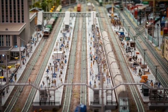 A detailed model of a bustling train station with multiple platforms and miniature figures of people. There are several tracks and model trains, along with buildings and station infrastructure such as clocks and lights. The setup is intricately crafted, displaying a scene of everyday life and transit.