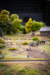 A miniature scene depicting a rural farming landscape with small human figures engaged in agricultural activities. Fields are being tended to, and there are tiny figures doing tasks like planting and carrying sacks. There are sheep grazing in a fenced area, a cart, and a horse. In the background, there's a small wooden structure resembling a barn or house, and lush green trees surround the area.