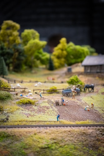 A miniature scene depicting a rural farming landscape with small human figures engaged in agricultural activities. Fields are being tended to, and there are tiny figures doing tasks like planting and carrying sacks. There are sheep grazing in a fenced area, a cart, and a horse. In the background, there's a small wooden structure resembling a barn or house, and lush green trees surround the area.