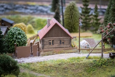 A small wooden house with a red roof is surrounded by a low wooden fence. Lush green trees and plants are in the background, with a gravel path leading up to the house. A miniature greenhouse and some gardening tools are visible nearby.