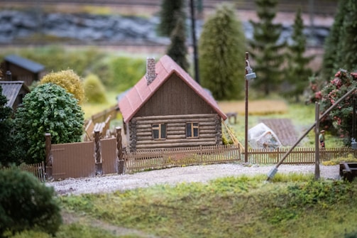 A small wooden house with a red roof is surrounded by a low wooden fence. Lush green trees and plants are in the background, with a gravel path leading up to the house. A miniature greenhouse and some gardening tools are visible nearby.