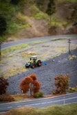 A model landscape with a small tractor plowing or working on a patch of dark brown soil. There are miniature trees with reddish-brown foliage, and a curved model road is visible in the foreground. The background shows a hill with sparse vegetation.