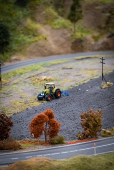 A model landscape with a small tractor plowing or working on a patch of dark brown soil. There are miniature trees with reddish-brown foliage, and a curved model road is visible in the foreground. The background shows a hill with sparse vegetation.