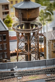 A model train set scene featuring a large, rusted water tower on an elevated metal structure. The area is surrounded by model buildings, fences, and a railroad track in the foreground, depicting an industrial or urban environment.