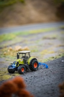 A miniature green tractor with large black tires is situated on a textured surface resembling a farm field. The tractor features a detailed design and is towing a blue agricultural implement. The background has a blurred effect with shades of green and brown, suggesting a natural, rural landscape.