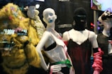 The image features a display of fashion mannequins adorned with eclectic and avant-garde outfits. The left mannequin is draped in a vibrant, textured yellow coat with butterfly embellishments. The center mannequin wears a black ski mask and a silky red dress with delicate lace, accented by bold, chunky accessories. The right mannequin showcases a modern outfit with exaggerated features, including a large bow hat. Neon reflections from a cityscape are visible in the background, adding an urban vibe.