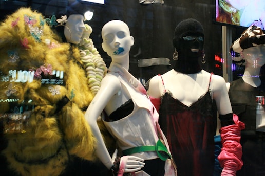 The image features a display of fashion mannequins adorned with eclectic and avant-garde outfits. The left mannequin is draped in a vibrant, textured yellow coat with butterfly embellishments. The center mannequin wears a black ski mask and a silky red dress with delicate lace, accented by bold, chunky accessories. The right mannequin showcases a modern outfit with exaggerated features, including a large bow hat. Neon reflections from a cityscape are visible in the background, adding an urban vibe.