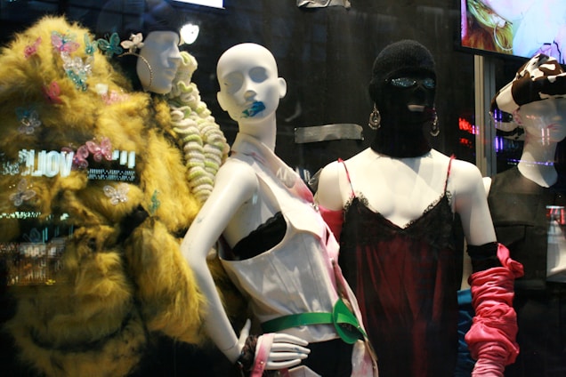 The image features a display of fashion mannequins adorned with eclectic and avant-garde outfits. The left mannequin is draped in a vibrant, textured yellow coat with butterfly embellishments. The center mannequin wears a black ski mask and a silky red dress with delicate lace, accented by bold, chunky accessories. The right mannequin showcases a modern outfit with exaggerated features, including a large bow hat. Neon reflections from a cityscape are visible in the background, adding an urban vibe.