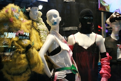 The image features a display of fashion mannequins adorned with eclectic and avant-garde outfits. The left mannequin is draped in a vibrant, textured yellow coat with butterfly embellishments. The center mannequin wears a black ski mask and a silky red dress with delicate lace, accented by bold, chunky accessories. The right mannequin showcases a modern outfit with exaggerated features, including a large bow hat. Neon reflections from a cityscape are visible in the background, adding an urban vibe.
