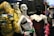 The image features a display of fashion mannequins adorned with eclectic and avant-garde outfits. The left mannequin is draped in a vibrant, textured yellow coat with butterfly embellishments. The center mannequin wears a black ski mask and a silky red dress with delicate lace, accented by bold, chunky accessories. The right mannequin showcases a modern outfit with exaggerated features, including a large bow hat. Neon reflections from a cityscape are visible in the background, adding an urban vibe.