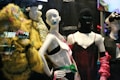 The image features a display of fashion mannequins adorned with eclectic and avant-garde outfits. The left mannequin is draped in a vibrant, textured yellow coat with butterfly embellishments. The center mannequin wears a black ski mask and a silky red dress with delicate lace, accented by bold, chunky accessories. The right mannequin showcases a modern outfit with exaggerated features, including a large bow hat. Neon reflections from a cityscape are visible in the background, adding an urban vibe.