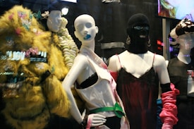 The image features a display of fashion mannequins adorned with eclectic and avant-garde outfits. The left mannequin is draped in a vibrant, textured yellow coat with butterfly embellishments. The center mannequin wears a black ski mask and a silky red dress with delicate lace, accented by bold, chunky accessories. The right mannequin showcases a modern outfit with exaggerated features, including a large bow hat. Neon reflections from a cityscape are visible in the background, adding an urban vibe.