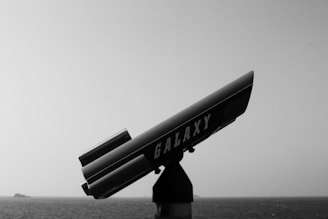 A black and white image of a telescope labeled 'Galaxy' mounted on a stand. The telescope is pointed towards the sea, with faint landmasses visible in the distance under a clear sky. The scene appears calm with minimal visual clutter.