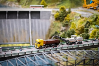 A detailed diorama of a construction scene on a bridge, featuring a yellow truck and a machinery vehicle loading gravel. Tiny model figures are positioned around the construction site, set against a backdrop of a dam and lush, green trees.