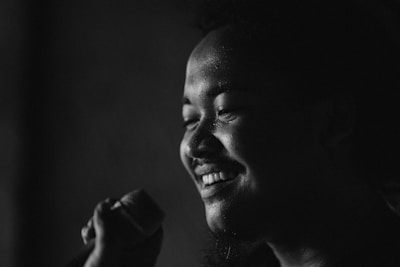 A person is holding a microphone and smiling. The image is in black and white, creating a striking contrast between the highlights on the person's face and the dark background.