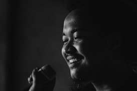 A person is holding a microphone and smiling. The image is in black and white, creating a striking contrast between the highlights on the person's face and the dark background.