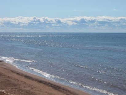 The image depicts a tranquil beach scene with gentle waves lapping at the sandy shore. The expansive blue ocean stretches out to the horizon, where it meets a sky filled with scattered, fluffy clouds. Sunlight glints off the water, creating a shimmering effect.